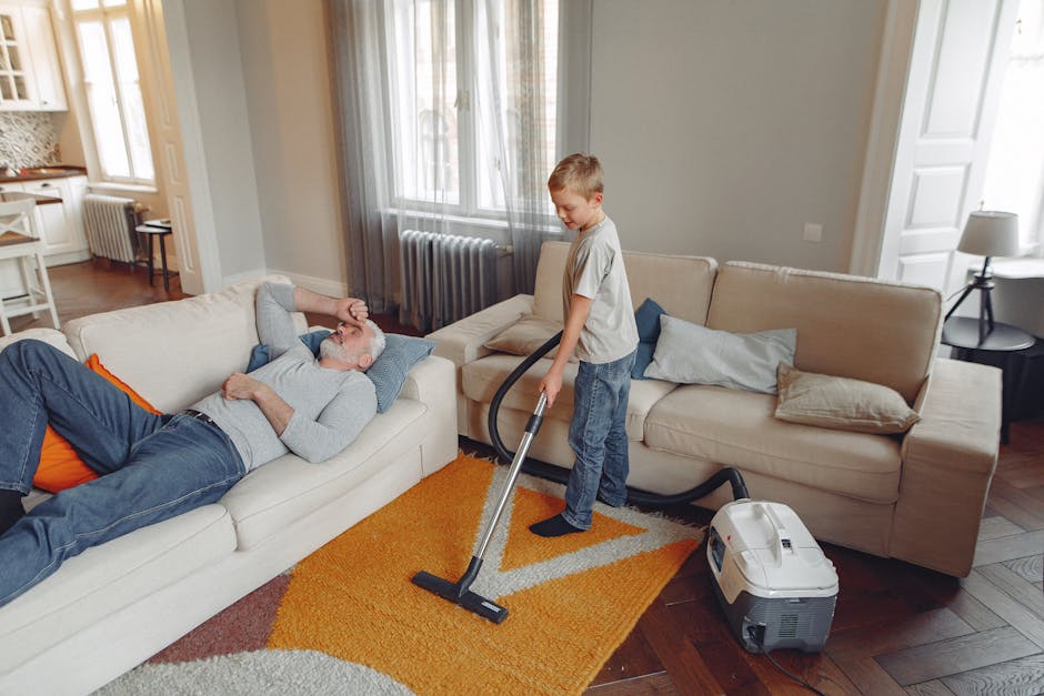 A young boy is vacuuming the living room carpet with a handheld vacuum cleaner, while an older man with gray hair and a beard is lying on a white sofa, resting with his eyes closed and one hand on his forehead. The living room features light gray walls, a large window with sheer curtains letting in natural light, and wooden flooring partially covered by an orange and cream area rug. The sofa set includes beige cushions, and a side table with a lamp is visible beside the sofa. The scene suggests routine domestic surface cleaning and maintenance by Belsize Park Carpet Cleaning, emphasizing a tidy and hygienic home environment.
