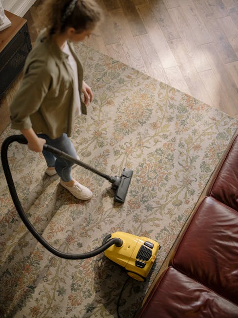 A woman using a yellow vacuum cleaner to perform surface cleaning and deep cleaning on a beige and floral patterned rug in a living room. The room features wooden flooring adjacent to the rug, a red leather sofa on one side, and natural light illuminating the space. The woman, dressed in casual attire with a beige jacket and white shoes, is focusing on removing dust and dirt from the rug. This image illustrates professional domestic cleaning services offered by Belsize Park Carpet Cleaning, showcasing the process of sanitisation and maintenance for home carpets in NW3, BELSIZE PARK.