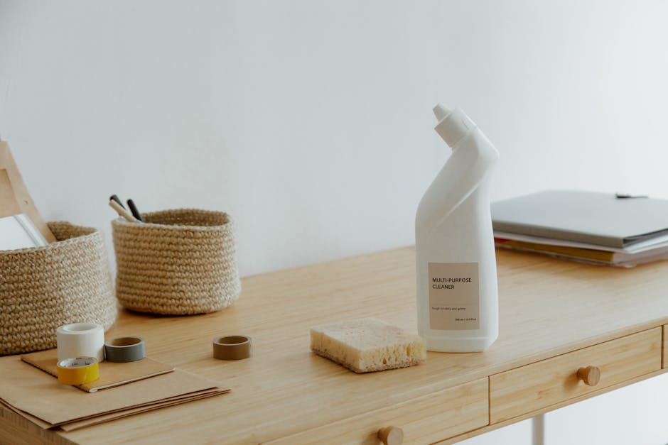 A wooden desk with a light finish, positioned against a plain white wall, displaying cleaning supplies and organisers. On the right side, there is a white multi-purpose cleaning spray bottle with a beige label, accompanied by a yellow and white sponge. To the left, two round woven baskets hold miscellaneous items such as pens or small accessories. Several small open containers in neutral tones are arranged on a piece of brown craft paper, likely for organizing small items. In the background, a closed laptop and a stack of papers are partially visible, indicating a workspace setup. The scene is well-lit with soft, natural lighting, emphasizing the tidy, clean surface suitable for surface cleaning or sanitisation, reflecting a professional approach to domestic or office cleaning by Belsize Park Carpet Cleaning.